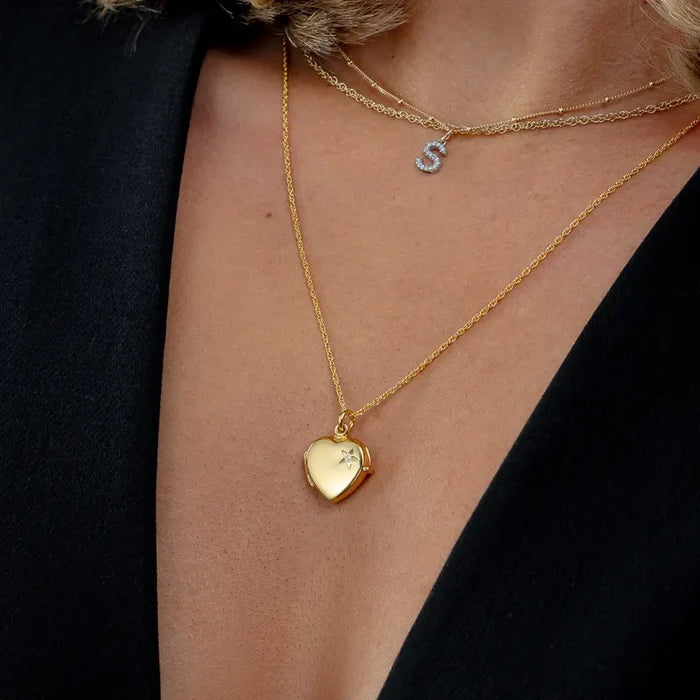 Close-up of a gold heart locket necklace with a diamond star detail, layered with fine gold chains and a diamond initial “S” pendant, styled against a black blazer.