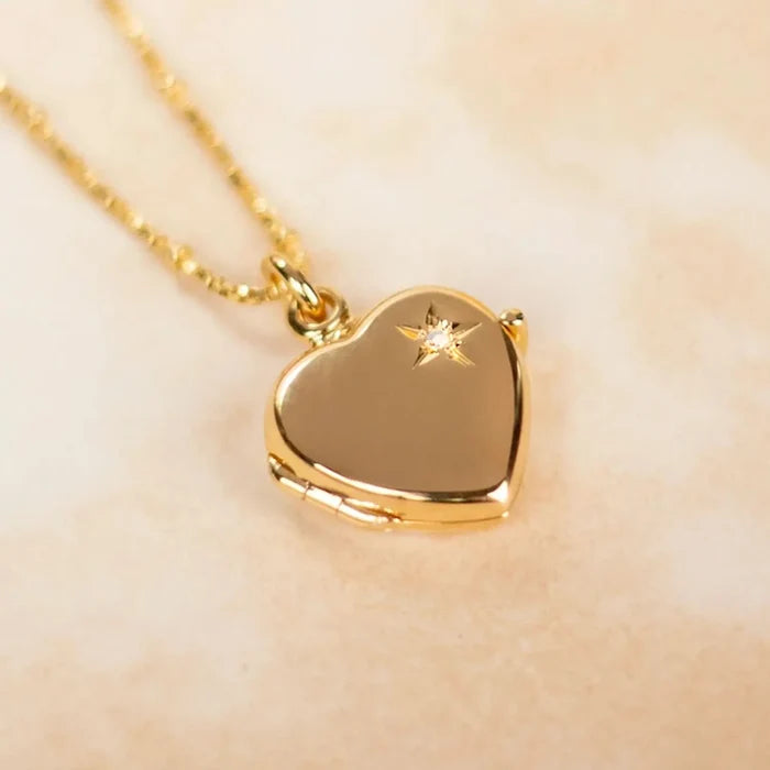 lifestyle image of a gold heart-shaped locket necklace, on a beaded chain featuring a delicate starburst engraving with a central diamond. Shown against a soft, neutral background.