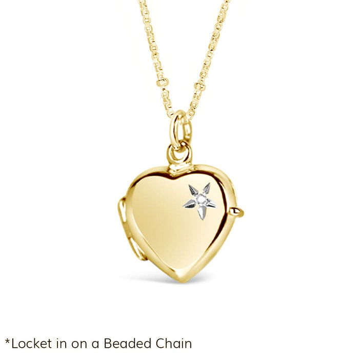 Studio product photo of a gold heart-shaped locket necklace with a diamond starburst engraving on the front. The locket hangs from a delicate beaded gold chain, displayed against a white background