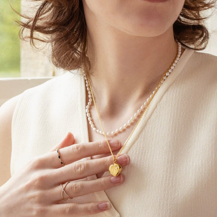 Woman in a sleeveless cream top, wearing a 2 photo diamond heart locket in gold and a pearl necklace, holding the gold locket