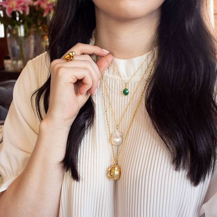 Ivory baroque pearl necklace layered with other gold necklaces. Woman is wearing a white textured top.