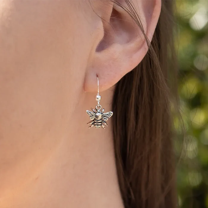Close-up of a sterling silver bee drop earring worn on the ear, showing the detailed bee charm suspended from a delicate hook fastening.
