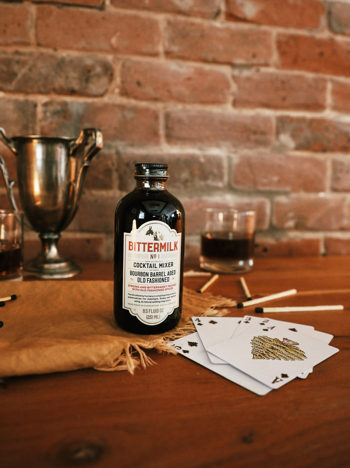 A bottle of Bittermilk Old Fashioned Mix sits on a wooden surface, in front of a brick wall, accompanies with playing cards, wooden matches, trophies.