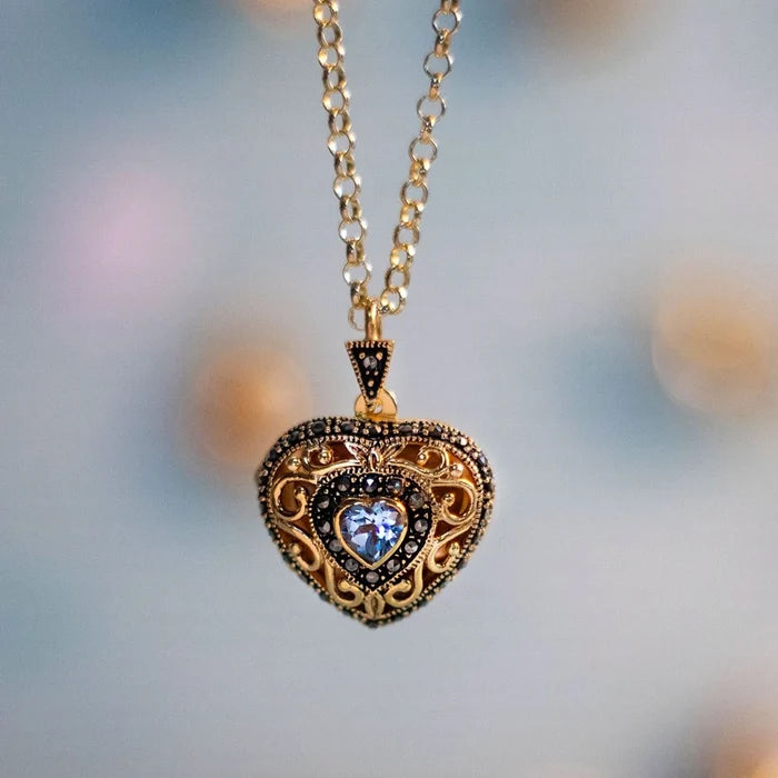 Gold heart shaped locket necklace n a circular linked gold chain. The locket has a detailed filigree design and features a blue topaz gemstone at it's centre