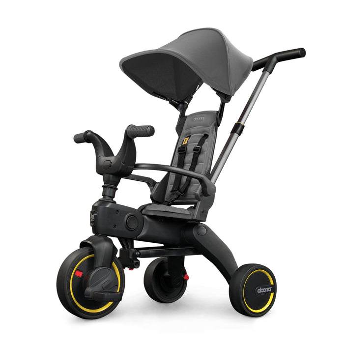 A compact grey Doona Liki Trike S1 with yellow accents, featuring a multi-position reclining backrest, parent control bar, footrest, armrest, and canopy.