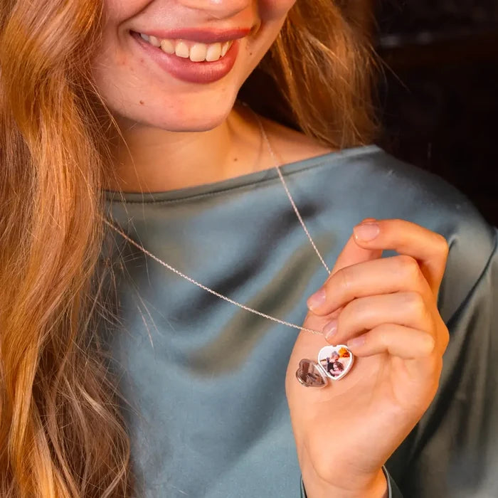 Close-up of a woman smiling while holding a heart-shaped silver locket open on a chain, revealing miniature photos inside.