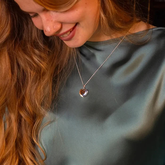 A woman in a teal satin blouse smiles as she looks down at her closed heart-shaped silver locket necklace.