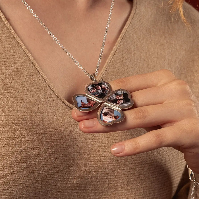 A close-up of a person wearing a silver necklace with a four photo pendant. The person is wearing a brown knit top, and the pendant is resting on their hand.