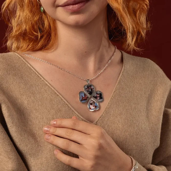 A close-up of a woman wearing a silver four-leaf clover locket necklace with custom photo compartments, styled with a beige V-neck sweater against a dark red background.