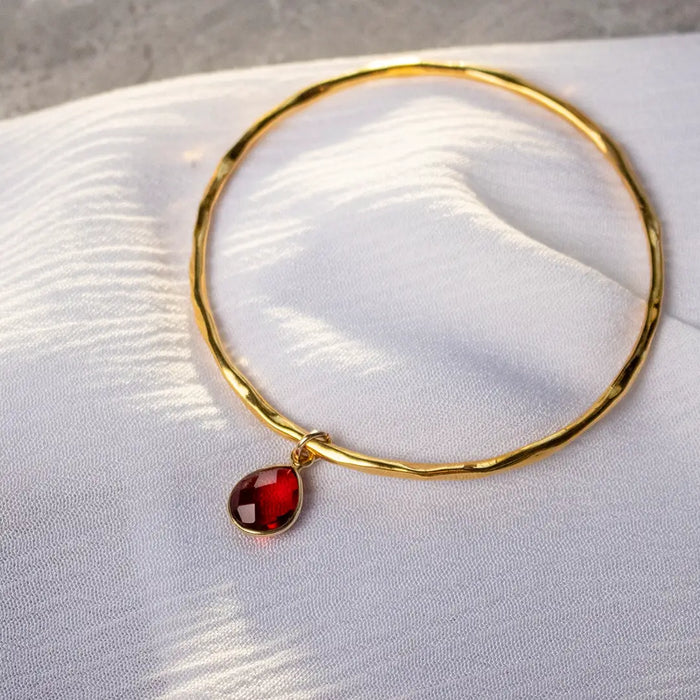 Gold hammered bangle with single garnet teardrop charm resting on white fabric, highlighting the rich red gemstone and polished finish