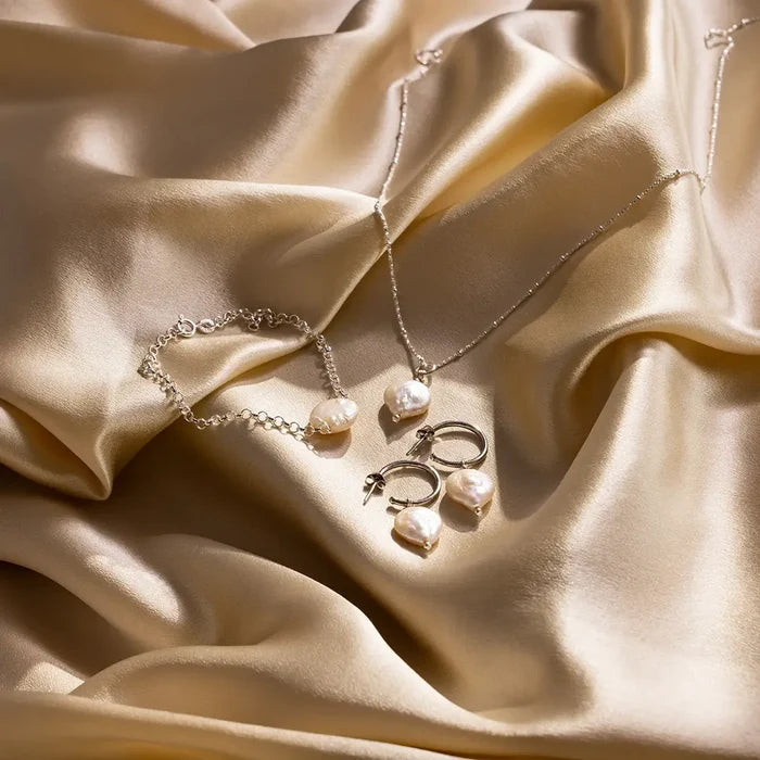 Sterling silver coin pearl jewellery set including bracelet, necklace and hoop earrings displayed on gold satin fabric, highlighting natural freshwater pearl texture