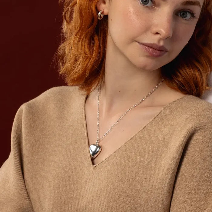 A close-up of a woman with red hair wearing a silver heart-shaped pendant necklace and a small hoop earring, dressed in a beige knit sweater against a dark red background.
