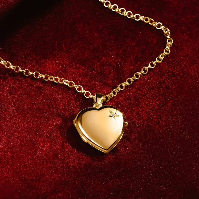 A gold heart-shaped locket necklace by Lily Blanche, with a small diamond on the front, displayed on a rich red velvet background. The locket hangs from a gold chain, catching the light with a polished shine.