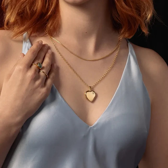 A close-up lifestyle shot of a model wearing a gold heart pendant necklace layered over a gold chain, paired with a light blue satin top.