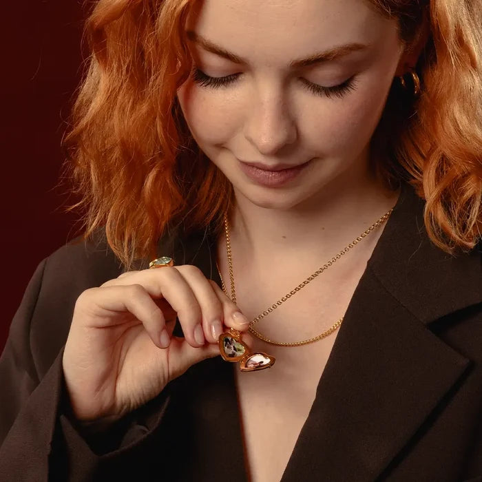 A close-up lifestyle shot of a woman with red hair wearing a delicate gold chain necklace with a heart-shaped pendant, open showing two photos inside, set against a dark brown blazer and a dark red background.