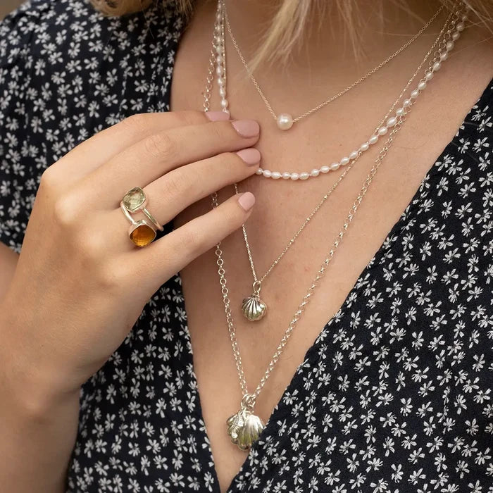 Close-up of layered sterling silver necklaces featuring a shell locket and pearl necklace, styled with gemstone rings and a floral dress.