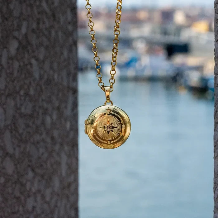 Gold compass locket necklace suspended outdoors between stone walls with soft blurred background