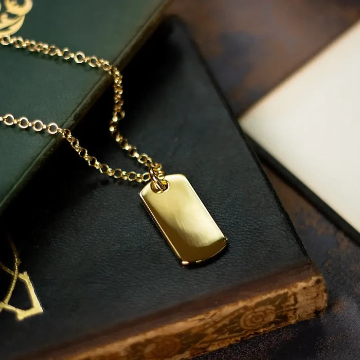 Gold dog tag pendant necklace resting on vintage books, highlighting the polished finish and curved rectangular shape