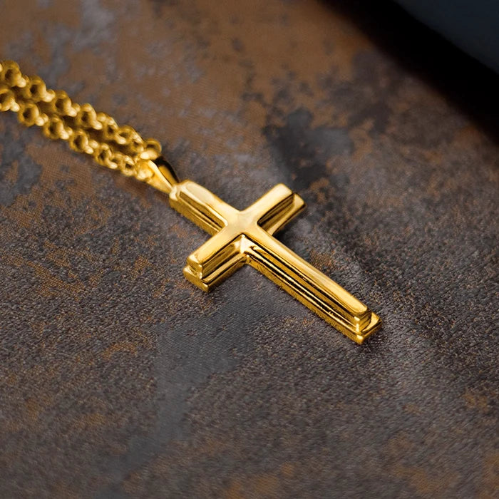 Gold cross pendant necklace by Lily Blanche on a chain, photographed close-up on a textured dark surface, highlighting the polished finish and clean, classic design.