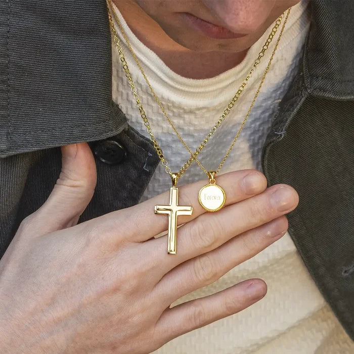 Close-up of a person wearing layered gold necklaces by Lily Blanche featuring a heavy gold cross pendant and a small round engraved disc reading “Forever”