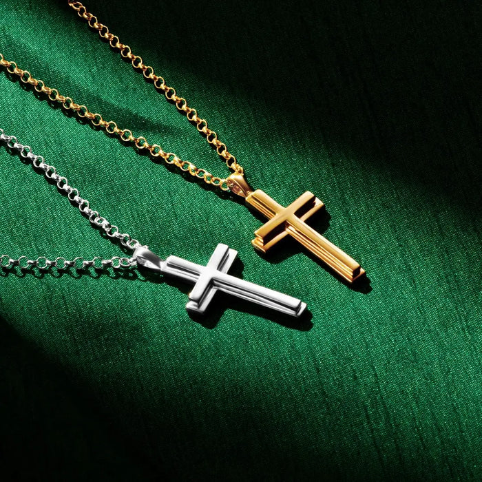 Two cross necklaces, one gold and one silver by Lily Blanche, displayed on a rich green fabric background. The gold cross hangs from a gold chain in the upper right, while the silver cross hangs from a silver chain below. Both pendants have a polished, bevelled design with dramatic lighting creating shadows on the fabric.