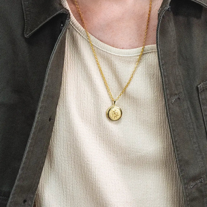Close-up of a person wearing a gold St Christopher locket pendant necklace on a belcher chain over a textured cream top
