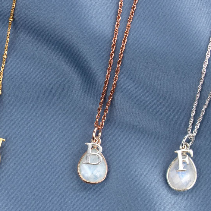 two moonstone charm necklaces in silver and rose gold  with silver initial charms photographed on blue satin fabric