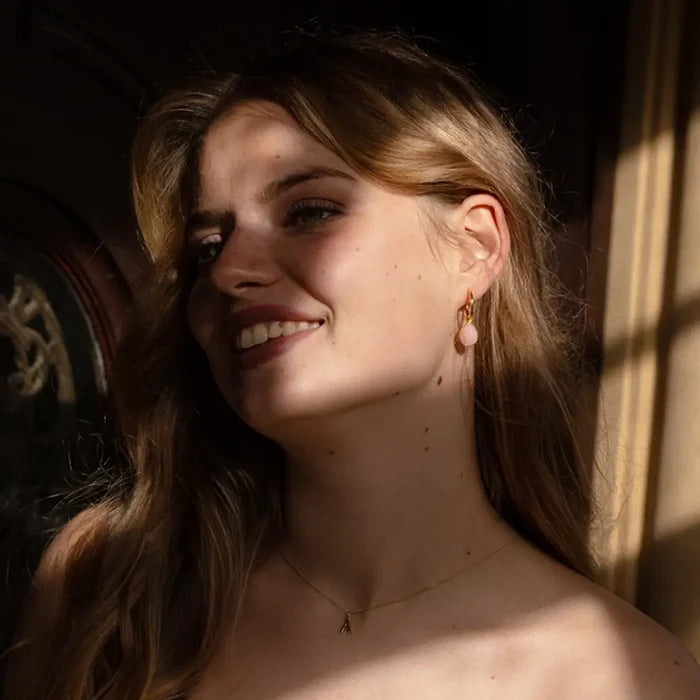 Woman smiling in soft sunlight, wearing gold hoop earrings with teardrop-shaped pink opal gemstones and a delicate gold initial necklace