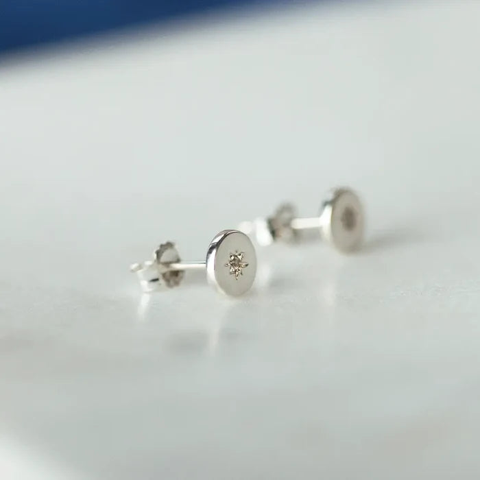 Sterling silver round stud earrings with small diamond star detail, displayed on a soft neutral surface with a minimalist aesthetic