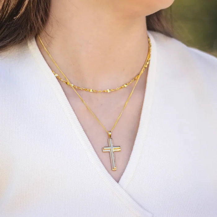 woman wearing a white v neck top with two layered gold necklaces. The shorter necklace is a decorative twist chain and the longer necklace is a simple chain with a large gold cross pendant by Lily Blanche.