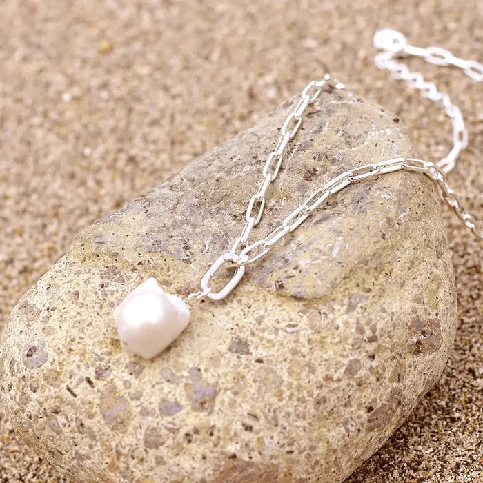 Sterling silver paperclip chain necklace with a single ivory baroque freshwater pearl pendant, displayed draped over a smooth beach stone on sand in natural sunlight.
