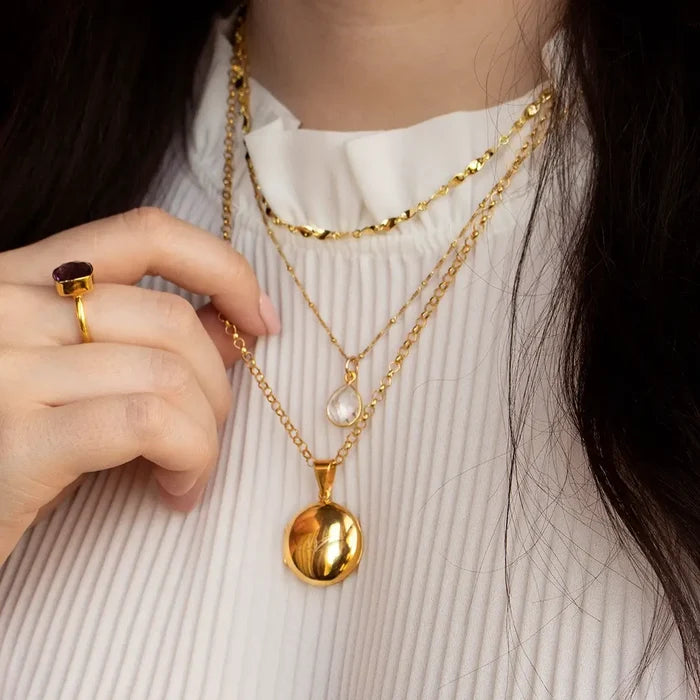 Layered gold necklaces worn with a gold locket pendant and white quartz charm necklace, styled with a gold gemstone ring and cream blouse.